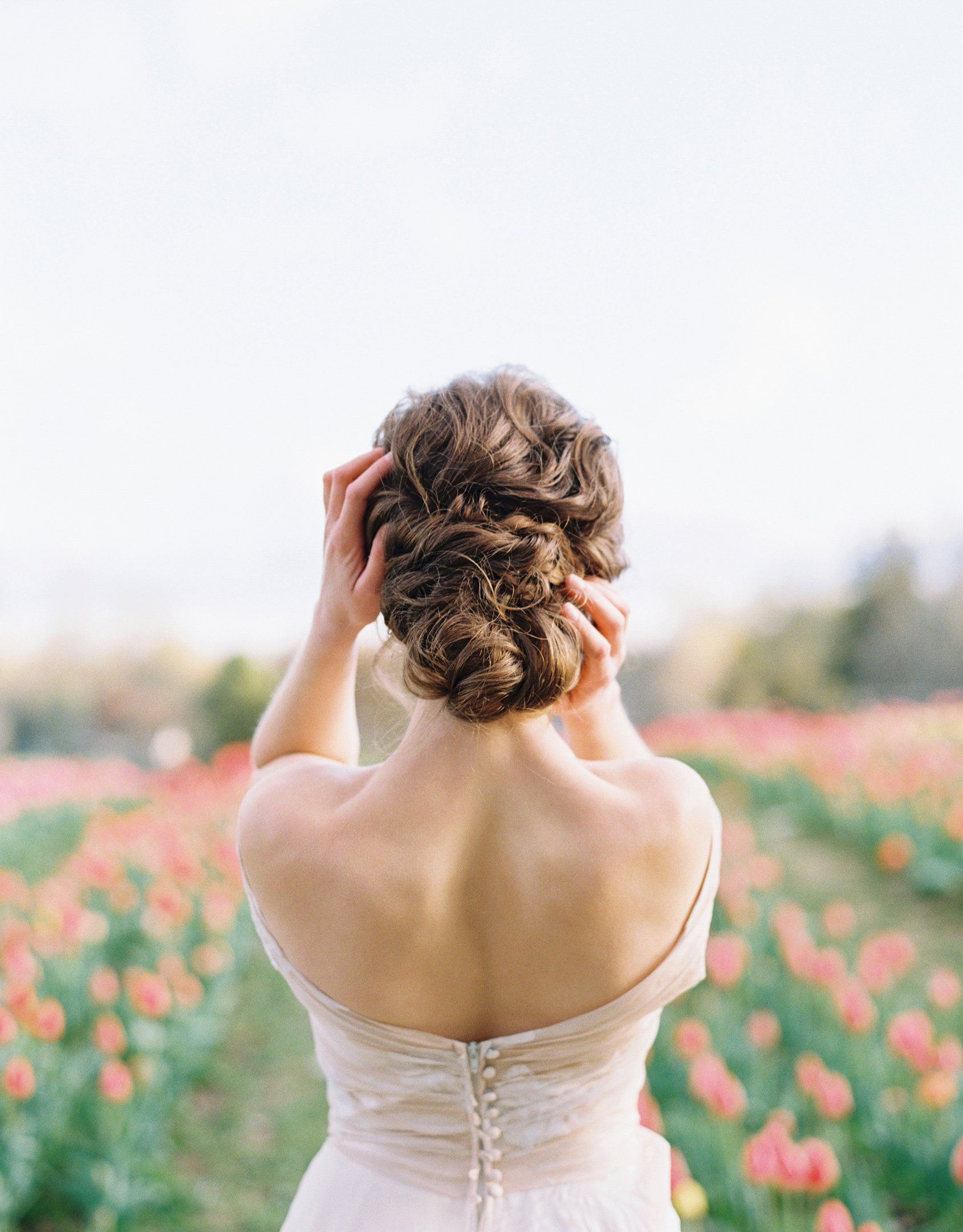 Bride in strapless gown with romantic updo hairstyle standing in tulip field, holding hair with both hands on wedding day.